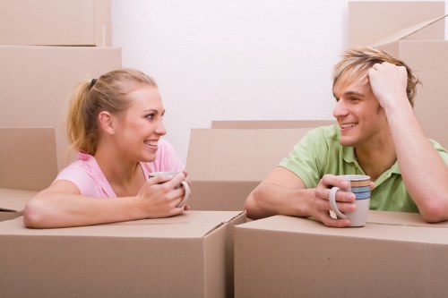 Two young adults, a blonde woman wearing a pink top and a blonde man in a green T-shirt, are sitting inside a room filled with stacked cardboard moving boxes. The woman is holding a smartphone, smiling, and looking at the man, who is seated with his left hand resting on his head and holding a striped coffee mug in his right hand. The background shows a plain white wall and additional cardboard boxes, indicating an ongoing household move, possibly around Clapham or the surrounding South London area. The boxes are of standard brown kraft cardboard, some with taped edges, and vary in size, suggesting they contain household personal belongings. The environment appears well-lit, clean, and organized, emphasizing a relaxed, casual atmosphere during a relocation process often managed by house removal services like Clapham Man with Van. The scene captures a moment of friendly interaction amid packing activities, typical of home removals in the Clapham district.