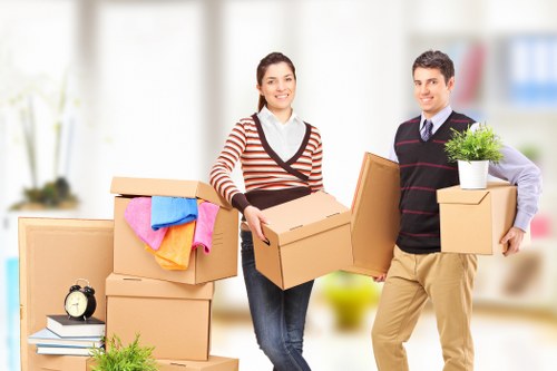 A man and a woman in a bright, modern residential interior surrounded by various packed cardboard boxes, some sealed with tape, and a few items such as a small black alarm clock on top of a box, a stack of books, and a potted plant. The woman, wearing a striped sweater, is holding an open cardboard box and smiling at the camera. The man, dressed in a sweater with a collared shirt underneath, is holding three boxes, one with a potted plant on top. The background shows a clean, well-lit living room with large windows, suggesting an active house removal or relocation scene, typical for a professional removals service like Clapham Man with Van in the Clapham area of London, sharing a tidy and organized environment suitable for house moves in the SW4 postcode area.