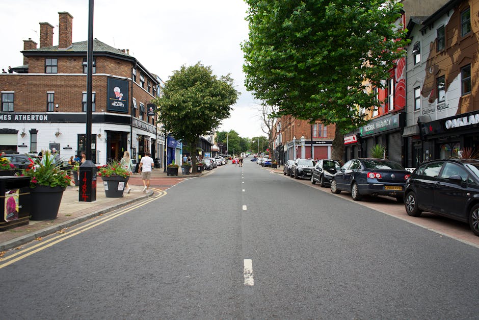 A straight urban street scene during daylight, featuring parked cars lining both sides of the road, with a variety of small shops and businesses occupying the ground floors of multi-storey brick build