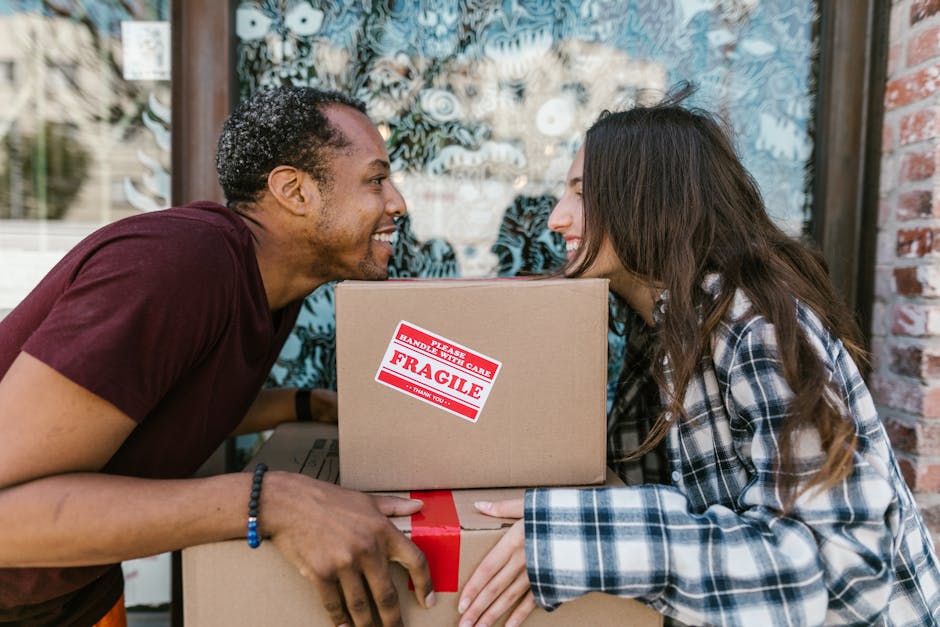 A young woman with curly black hair, wearing a white top, blue jeans, and white sneakers, is sitting on the wooden floor inside a room surrounded by numerous cardboard boxes used for home relocation. 